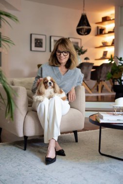 Çekici, orta yaşlı bir kadın sevimli köpeğiyle evdeki koltukta rahatlıyor. Süveter ve pantolon giyen sarışın kadın. Tam boy çekim.  