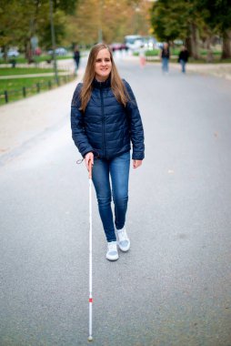 Beyaz bastonuyla parkta yürüyen görme engelli bir kadın. Sarı saçlı, sıradan kıyafetler giyen bir kadın. Tam boy çekim. 