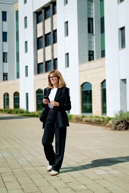 Şehir caddesinde yürüyen ve akıllı telefonunu kullanan orta yaşlı bir iş kadını. Sarı saçlı, siyah takım elbiseli ve gülümseyen bir kadın. Tam boy çekim. 