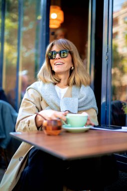 Orta yaşlı bir kadın kafenin dışında bir masada oturup kahvesini içiyor. Gündelik kıyafetler ve güneş gözlüğü takan kendine güvenen bir kadın..  