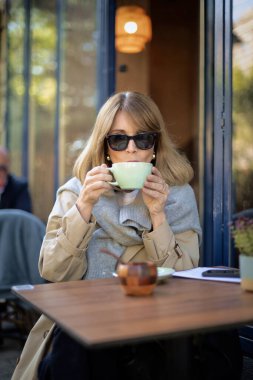 Orta yaşlı bir kadın kafenin dışında bir masada oturup kahvesini içiyor. Gündelik kıyafetler ve güneş gözlüğü takan kendine güvenen bir kadın.. 
