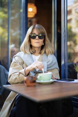Orta yaşlı bir kadın kafenin dışında bir masada oturup kahvesini içiyor. Gündelik kıyafetler ve güneş gözlüğü takan kendine güvenen bir kadın.. 