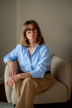 Sarışın, beyaz, koltukta dinlenen bir kadın. Mavi gömlek ve deve pantolonu giyen kendine güvenen bir kadın.. 