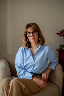 Sarışın, beyaz, koltukta dinlenen bir kadın. Mavi gömlek ve deve pantolonu giyen kendine güvenen bir kadın.. 