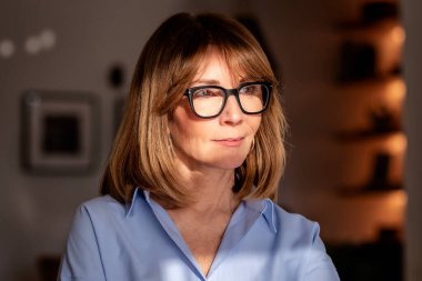 Samimi dairesinde duran gülümseyen kadının portresi. Mavi gömlek ve gözlük takan orta yaşlı kadın..