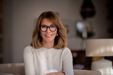 Çekici sarı saçlı kadın evdeki koltukta dinleniyor. Beyaz kazak giyen ve kameraya gülümseyen kendine güvenen bir kadın.. 