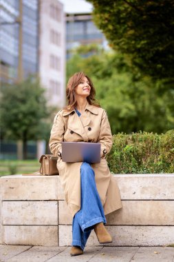 Esmer kadın şehir parkında oturuyor ve iş için dizüstü bilgisayar kullanıyor. Sıradan kıyafetler giyen mutlu orta yaşlı kadın. Tam boy çekim. 