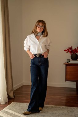 Oturma odasında duran sarı saçlı çekici bir kadının boyu. Beyaz gömlek ve kot pantolon giyen kendine güvenen bir kadın. Tam boy çekim. 