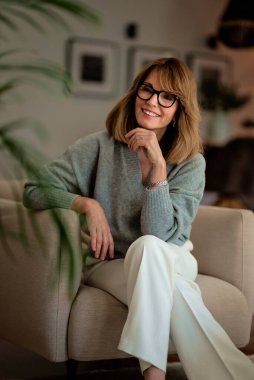 Çekici orta yaşlı kadın evdeki koltukta dinleniyor. Sarı saçlı, süveter ve pantolon giyen bir kadın..