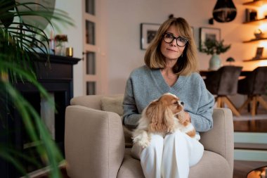 Çekici, orta yaşlı bir kadın sevimli köpeğiyle evdeki koltukta dinleniyor. Sarı saçlı, süveter ve pantolon giyen bir kadın..  