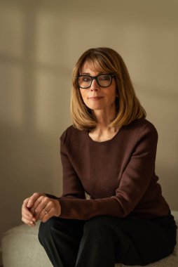 Duvarın önündeki taburede oturan gülümseyen kadın. Süveter ve pantolon giyen çekici bir kadın.. 