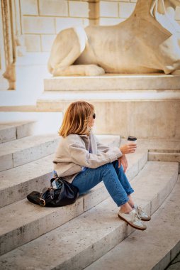Şehirdeki merdivenlerde oturan orta yaşlı bir kadın kahve içiyor. Tam yol ileri. Sarı saçlı kadın ceket ve kot pantolon giyiyor..  