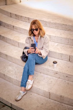 Şehirdeki bir merdivende oturan ve akıllı telefonunu kullanan orta yaşlı bir kadın. Tam yol ileri. Sarı saçlı kadın ceket ve kot pantolon giyiyor.. 
