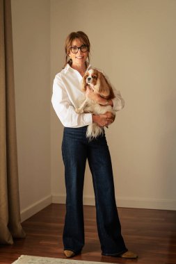 Orta yaşlı bir kadının sevimli köpeğiyle duvarın önünde duruşu. Beyaz gömlek ve kot pantolon giyen çekici bir kadın ve neşeli bir gülümseme.. 