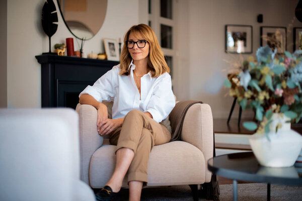 Smiling blond haired woman relaxing in an armchair in her cozy home. Attractive female wearing white shirt and pants and smiling.  