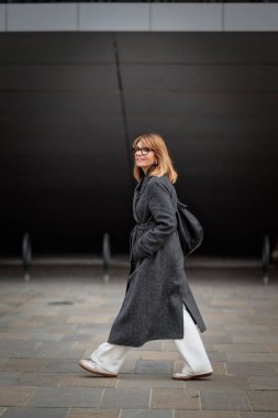 Uzun boylu, sarışın, olgun bir kadının sokakta yürümesi. Ceket ve beyaz pantolon giyen çekici bir kadın..  