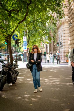 Şehir caddesinde yürüyen orta yaşlı bir kadın. Sarı saçlı, siyah ceketli, kot pantolonlu ve gülümseyen bir kadın..  