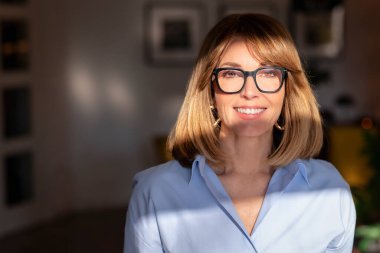 Sarı saçlı bir kadının, kameraya bakıp neşeli gülümsemesinin fotoğrafı. Mavi gömlekli ve okuma gözlüklü çekici bir kadın..  