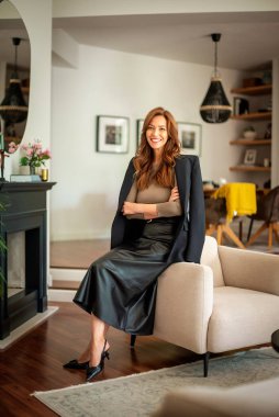 Çekici esmer saçlı kadın modern dairesinde koltukta dinleniyor. Orta yaşlı bir kadın kameraya bakıyor ve gülümsüyor. Tam boy çekim.  
