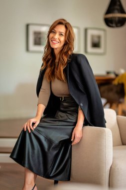 Çekici esmer saçlı kadın modern dairesinde koltukta dinleniyor. Orta yaşlı bir kadın kameraya bakıyor ve gülümsüyor..  