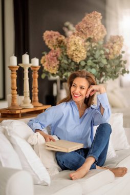 Güzel bir kadın evde rahatlatıcı bir günün tadını çıkarıyor. Orta yaşlı kadın rahat kanepesinde uzanmış kitap okurken ışıl ışıl gülümsüyordu. Tam uzunluk.  