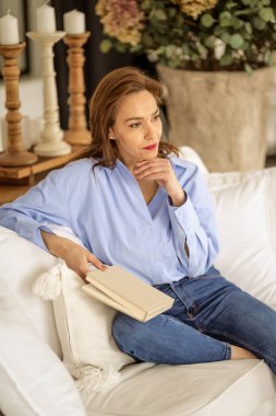 Güzel bir kadın evde rahatlatıcı bir günün tadını çıkarıyor. Orta yaşlı bir kadın, rahat kanepesinde uzanmış kitap okurken ışıl ışıl gülümserdi..  