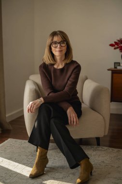 Oturma odasında rahat bir koltukta oturan ve kameraya gülümseyen olgun bir kadın. Süveter ve pantolon giyen sarışın kadın. Tam boy çekim.  