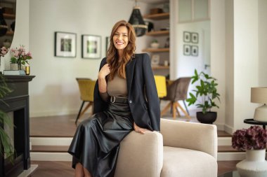 Oturma odasında rahat bir koltukta otururken gülümseyen olgun bir kadının portresi. Kumral saçlı, ceketli ve etekli çekici bir kadın..  