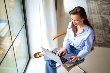 Pencerenin yanındaki sandalyede oturan çekici olgun kadın not defterini iş için kullanıyor. Beyaz kadın, evden çalışıyor. Merkez Ofis. 