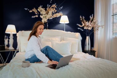 Evdeki yatakta oturan güzel bir kadın. Orta yaşlı kadın, rahat yatağında oturup internette gezinirken ışıl ışıl gülümsüyor. Merkez Ofis.  