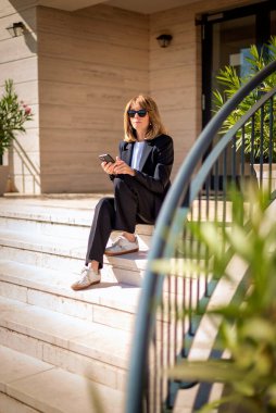 Evin girişindeki merdivenlerde oturan ve birini bekleyen orta yaşlı bir kadın. Cep telefonunu kullanan sarı saçlı bir kadın. Takım elbiseli bir iş kadını. Tam boy çekim.  