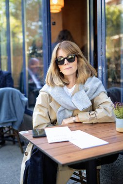 Kafenin dışındaki masada oturan orta yaşlı bir kadın. Sarı saçlı, günlük kıyafetler ve güneş gözlüğü takan bir erkek..  