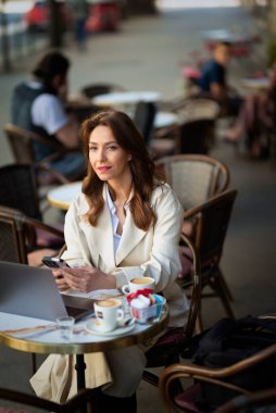 Masada oturan neşeli bir kadın, dizüstü bilgisayarla çalışıyor ve açık bir kafede akıllı telefon kullanıyor. Trençkot giyen çekici bir kadın..  