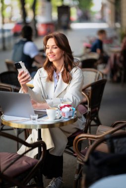 Masada oturan neşeli bir kadın, dizüstü bilgisayarla çalışıyor ve açık bir kafede akıllı telefon kullanıyor. Trençkot giyen çekici bir kadın..  