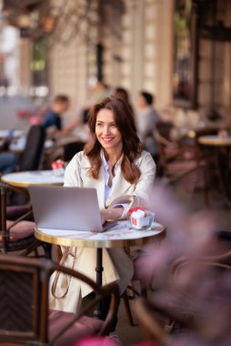 Neşeli bir kadın bir masada oturuyor, bir dizüstü bilgisayarla çalışıyor ve açık bir kafede bir fincan kahvenin tadını çıkarırken gülümsüyor. Trençkot giyen çekici bir kadın..  