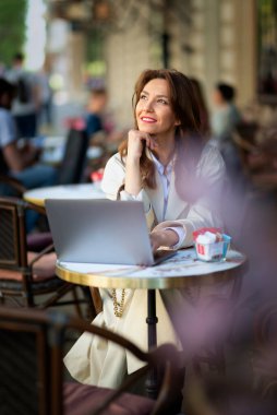 Neşeli bir kadın bir masada oturuyor, bir dizüstü bilgisayarla çalışıyor ve açık bir kafede bir fincan kahvenin tadını çıkarırken gülümsüyor. Trençkot giyen çekici bir kadın..  