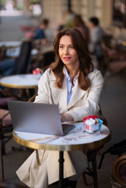Neşeli bir kadın bir masada oturuyor, bir dizüstü bilgisayarla çalışıyor ve açık bir kafede bir fincan kahvenin tadını çıkarırken gülümsüyor. Trençkot giyen çekici bir kadın..  