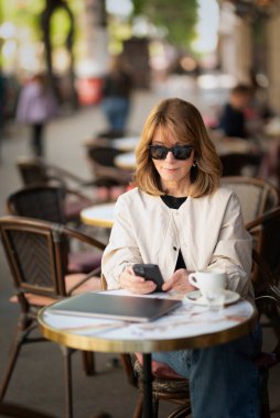 Orta yaşlı bir kadın kaldırımdaki kafede otururken mesaj atıyor. Sarı saçlı, güneş gözlüğü takan ve günlük kıyafetler giyen bir kadın..  