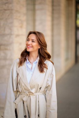 Güneşli bir günde, uzun saçlı, kendinden emin bir kadın şehir caddesinde yürüyor. Trençkot giyen ve neşeli gülümseyen çekici bir kadın..  