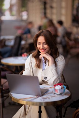 Neşeli bir kadın bir masada oturuyor, bir dizüstü bilgisayarla çalışıyor ve açık bir kafede bir fincan kahvenin tadını çıkarırken gülümsüyor. Trençkot giyen çekici bir kadın..  