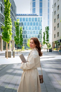 Kendine güvenen bir iş kadını elinde dizüstü bilgisayarla iş bölgesinde dolaşıyor. Trençkot giyen ve kameraya bakan orta yaşlı profesyonel bir kadın.. 