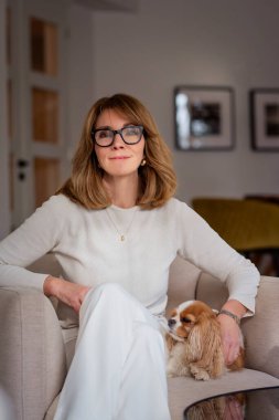 Sarışın bir kadın, evinde bir koltukta köpeğiyle oturuyor. Olgun kadın beyaz kazak ve pantolon giyiyor..  