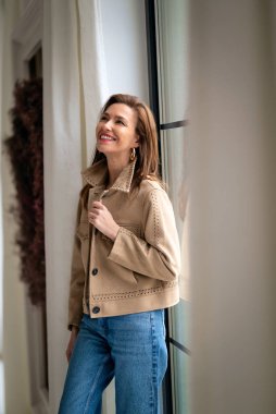 Esmer saçlı kadın pencerenin yanında duvara dayanıyor. Ceket giyen ve gülümseyen çekici bir kadın.. 