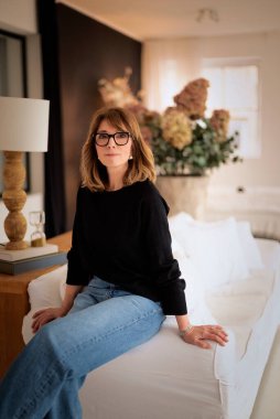 Orta yaşlı kadın gündelik kıyafetler giyip oturma odasındaki kanepede rahat bir pozisyonda oturuyor. Sarı saçlı kadın kameraya bakıyor ve gülümsüyor.. 