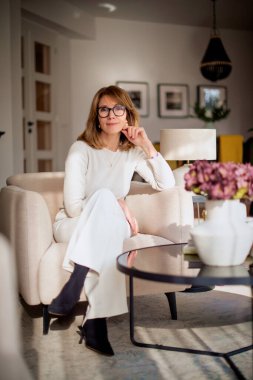 Zarif kıyafetler giymiş çekici bir kadın, evinin oturma odasındaki modern koltuğunda oturuyor. Tam boy çekim. 
