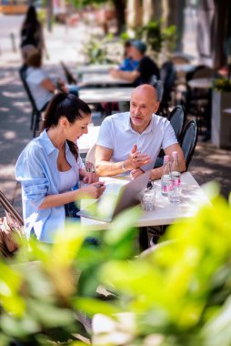  İnsanlar dışarıda bir toplantıda konuşuyorlar. Esmer kadın ve orta yaşlı adam kaldırımdaki kafede oturuyor. . 