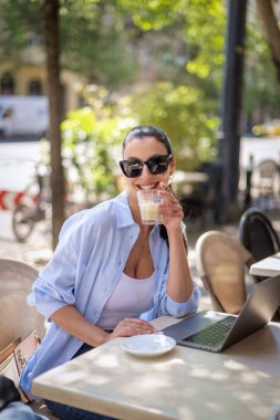 Esmer kadın bir masada oturuyor, açık hava kafesinde dizüstü bilgisayarla çalışıyor. Gündelik kıyafetler ve güneş gözlüğü takan çekici bir kadın..  