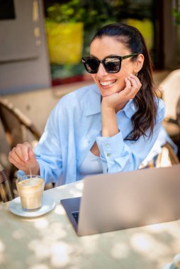 Esmer kadın bir masada oturuyor, açık hava kafesinde dizüstü bilgisayarla çalışıyor. Gündelik kıyafetler ve güneş gözlüğü takan çekici bir kadın..  
