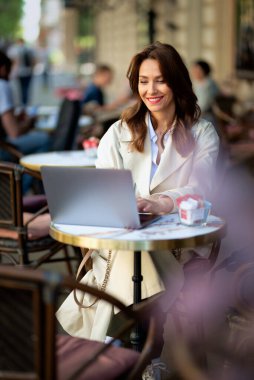 Masada oturmuş dışarıda bir kafede dizüstü bilgisayarla çalışan neşeli bir kadın. Trençkot giyen çekici bir kadın. Kendine güvenen profesyonel kadın video görüşmesi yapıyor..  