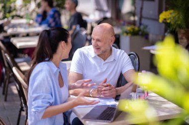  İnsanlar dışarıda bir toplantıda konuşuyorlar. Esmer kadın ve orta yaşlı adam kaldırımdaki kafede oturuyor.  